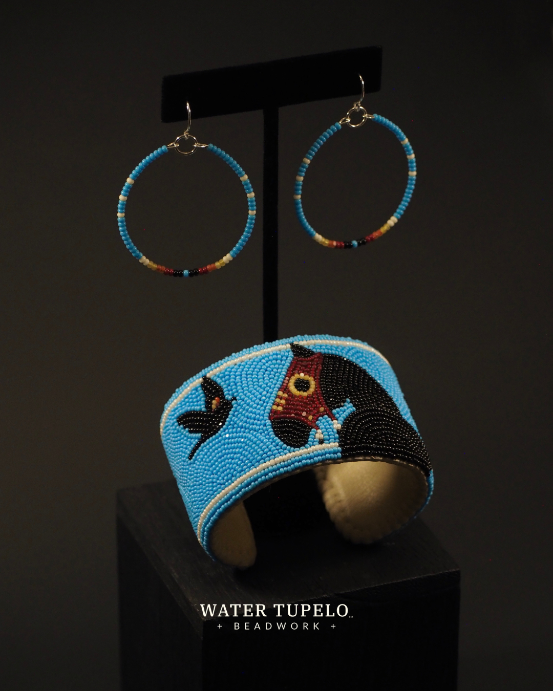 A jewelry set displayed on a black stand against a dark background, featuring Native American made turquoise beaded cuff with horse and red winged black bird beadwork and matching turquoise hoop earrings with sterling silver wire hooks. The brand name 'Water Tupelo Beadwork' is printed on the stand.