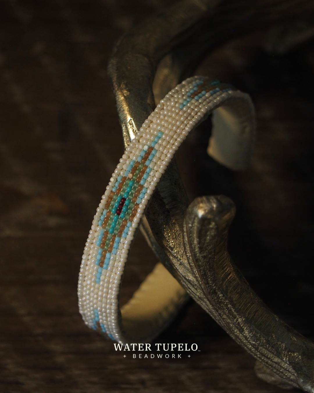 A beaded cuff bracelet with a Native American pattern in turquoise, brown, and white, displayed on a dark metal stand over a dark wooden surface. The brand name 'Water Tupelo Beadwork' is visible at the bottom.